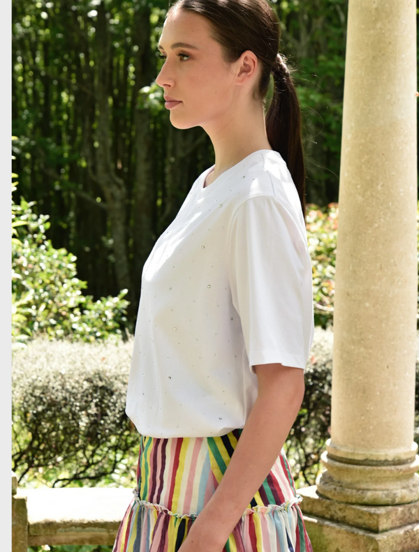 A woman in a white Sparkle Magic T-shirt paired with a vibrant multicolored striped skirt poses cheerfully outdoors.