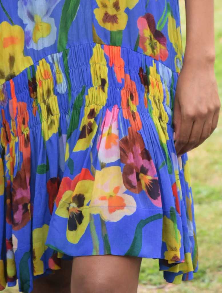 A woman in a cobalt floral dress poses beside a tree, emphasizing her stylish look in a natural setting.
