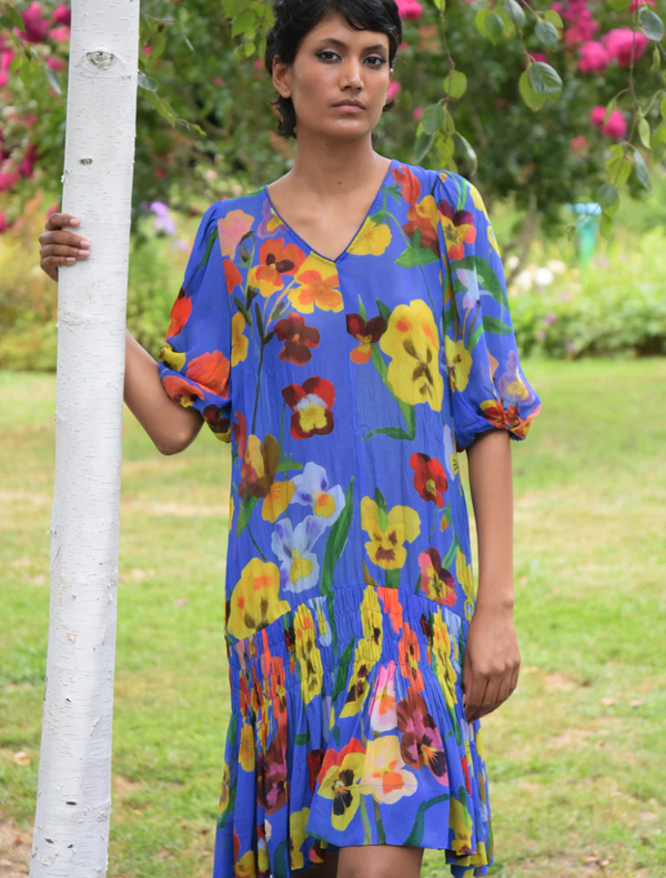 A woman in a cobalt floral dress stands beside a tree, showcasing her vibrant outfit against the natural backdrop.