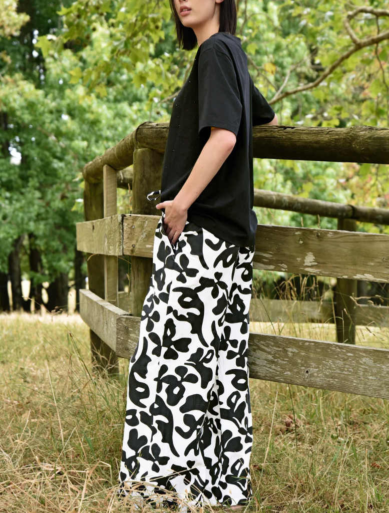 A woman in stylish black and white pants leans against a rustic fence, appearing calm and contemplative.