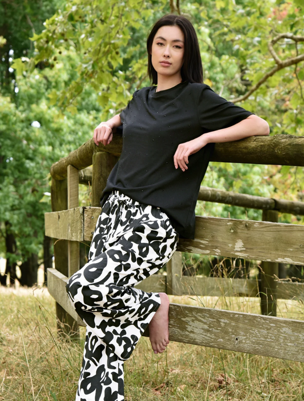 A woman in black and white pants leans against a wooden fence, looking relaxed and casual.