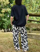 A woman wearing black and white pants leans casually against a fence, showcasing a relaxed outdoor atmosphere.