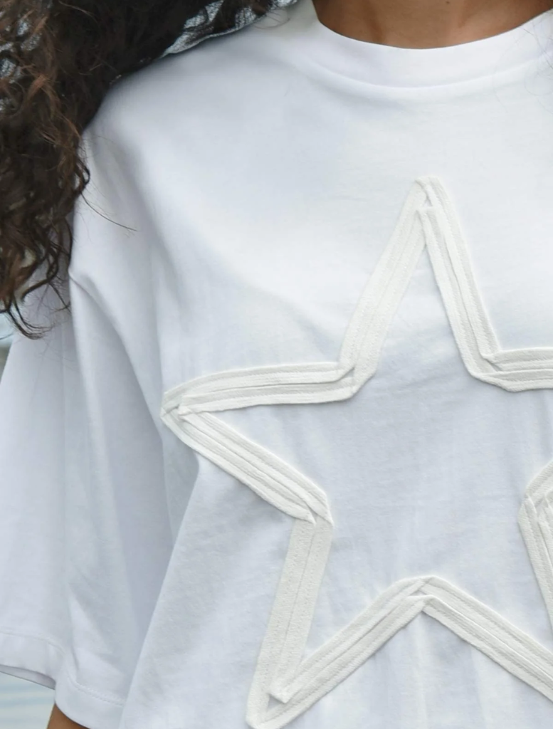 A woman in a white t-shirt displaying a star, standing outdoors with a soft-focus background.