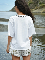 A woman dressed in a white shirt and shorts stands at the water's edge, looking out over the tranquil surface.
