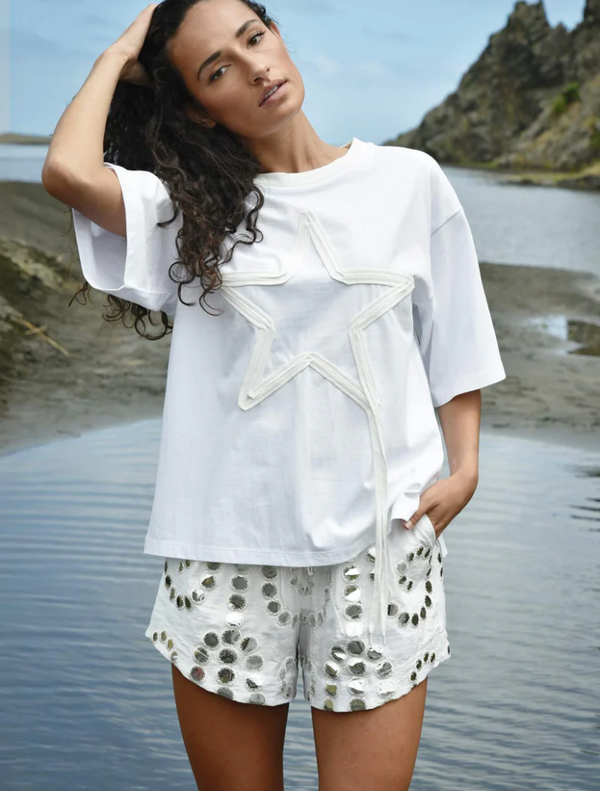 A woman in a white shirt and shorts stands by the water, gazing at the serene landscape around her.