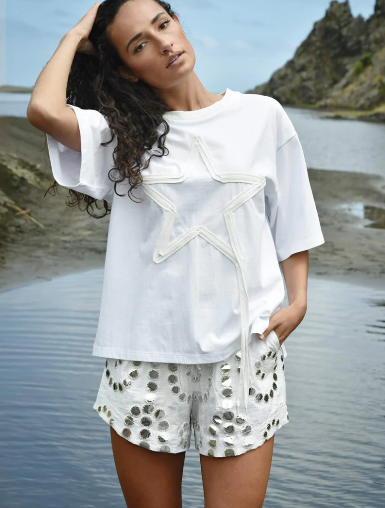 A woman in a white shirt and shorts stands by the water, gazing at the serene landscape around her.