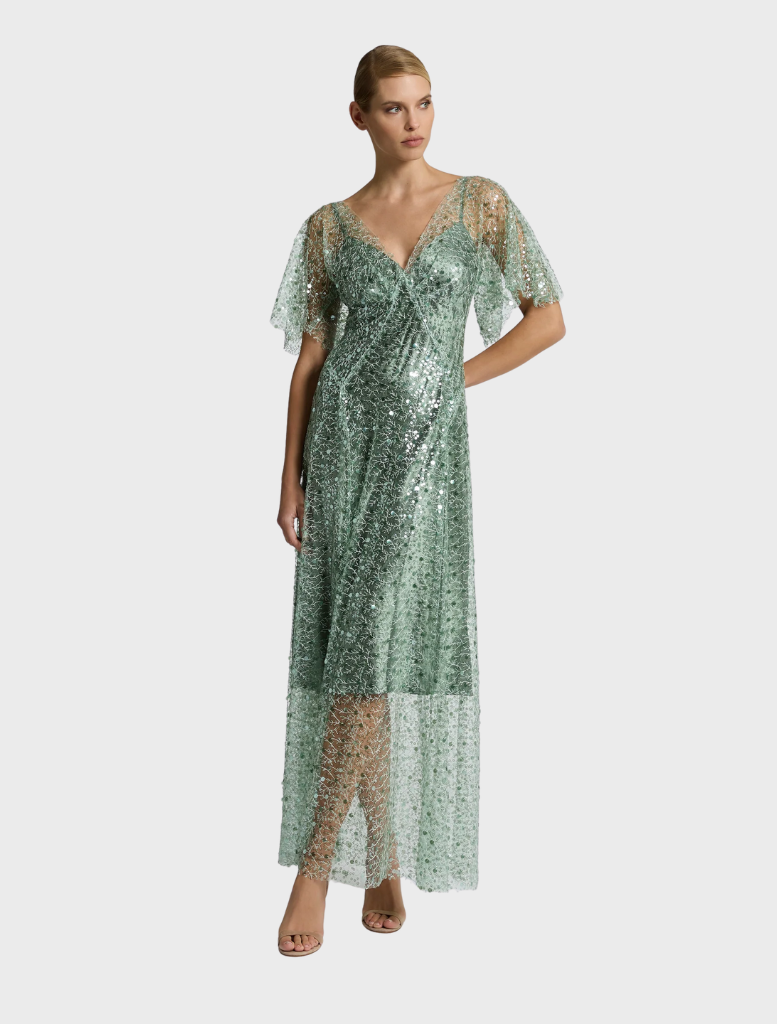 In a garden, a woman in a flowing sage Tilda gown stands amidst colorful flowers, embodying elegance and tranquility.