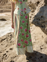 A woman in a floral dress stands on a rocky beach, wearing the Tillla Denim Maxi Skirt in Meadow Multi.