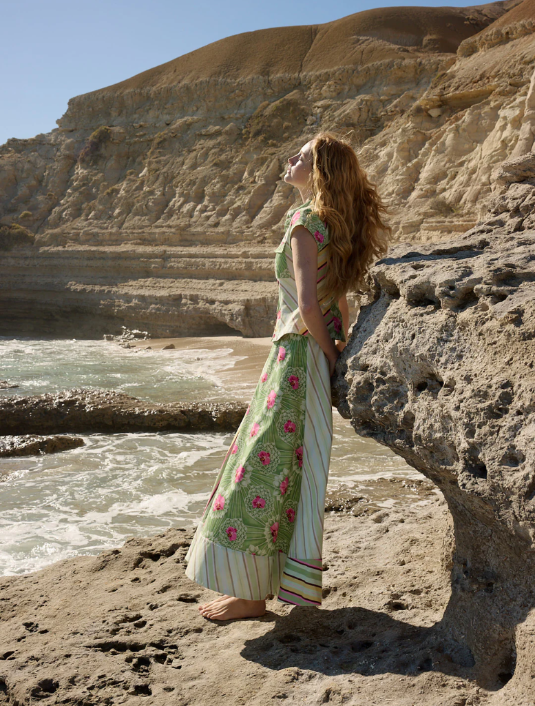 A woman in a floral dress, Tilla Denom Top - Meadow Multi, poses on a beach, surrounded by sand and ocean waves.