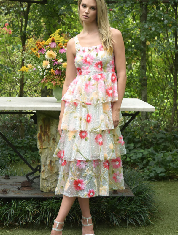 A woman in a floral dress stands gracefully in front of a vibrant garden filled with blooming flowers.