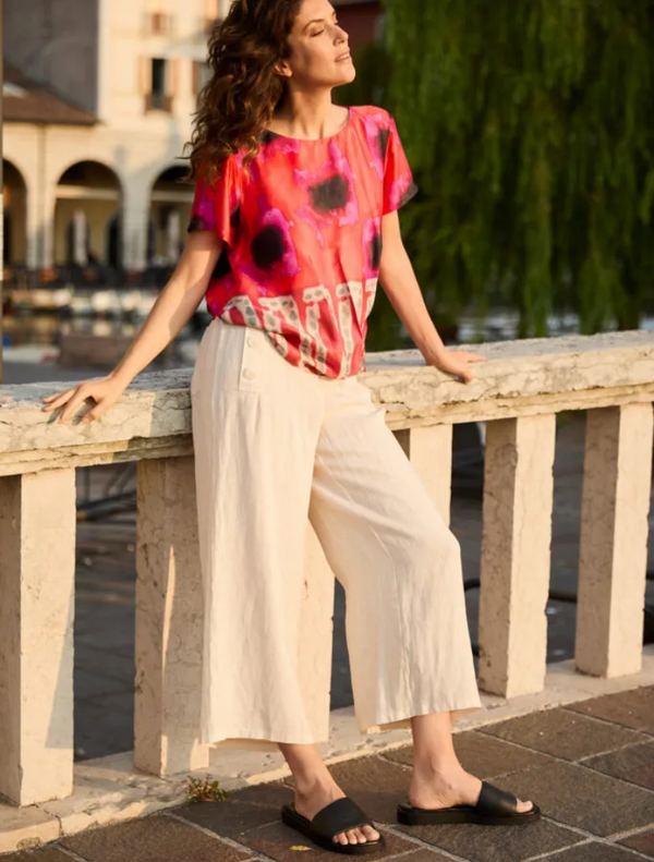 A woman dressed in a bright, patterned top and white pants poses, highlighting her stylish and colorful fashion choice.