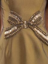 The Venus Midi Dress in gold showcases an off-the-shoulder style, embellished bow, fitted bodice, and a pencil skirt with a back split.