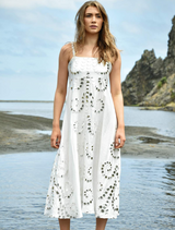 A woman wearing a white dress stands on the sandy beach, gazing at the ocean under a clear blue sky.