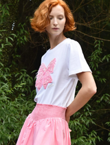  A woman wears a pink skirt and a white t-shirt with a bow, posing confidently in a sunny setting.