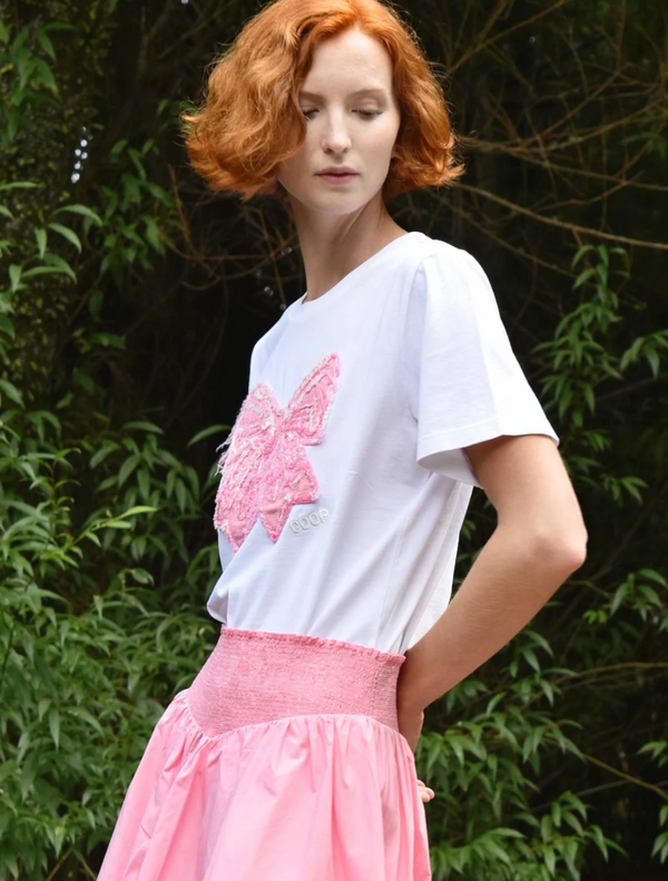  A woman wears a pink skirt and a white t-shirt with a bow, posing confidently in a sunny setting.