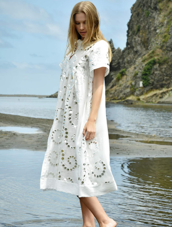 A woman wearing a white dress stands on the sandy beach, gazing at the ocean under a clear blue sky.