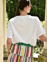 A woman wearing a white Sparkle Magic T-shirt and a striped skirt stands confidently, showcasing her stylish outfit.
