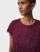 A woman in a burgundy fringed top stands confidently, showcasing her stylish outfit against a neutral background.