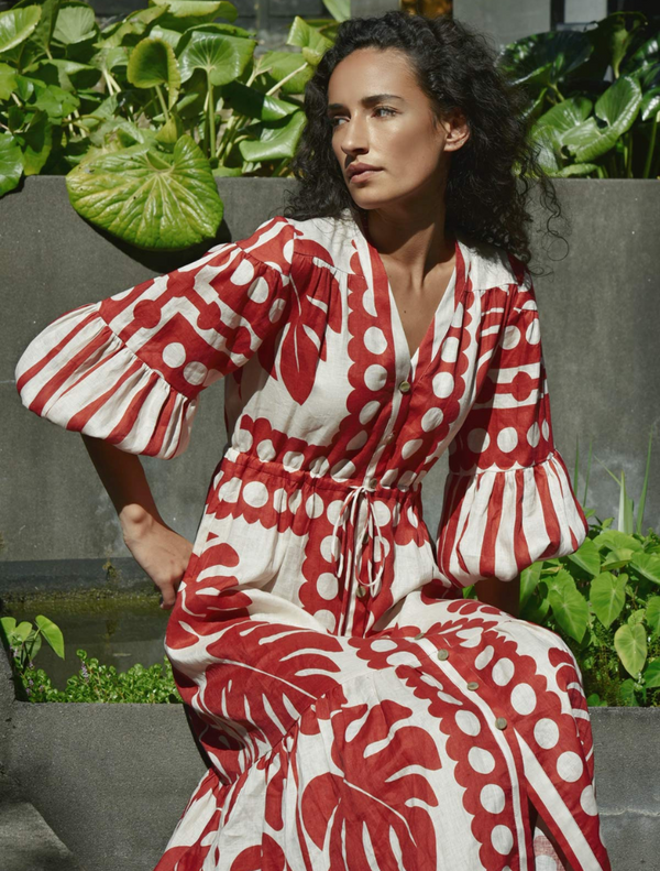 A woman in a stylish red and white dress stands before a wall, showcasing her outfit with a gentle smile.