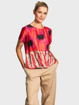 A woman in a lively, colorful top and white trousers stands, exuding a sense of style and confidence in her outfit.