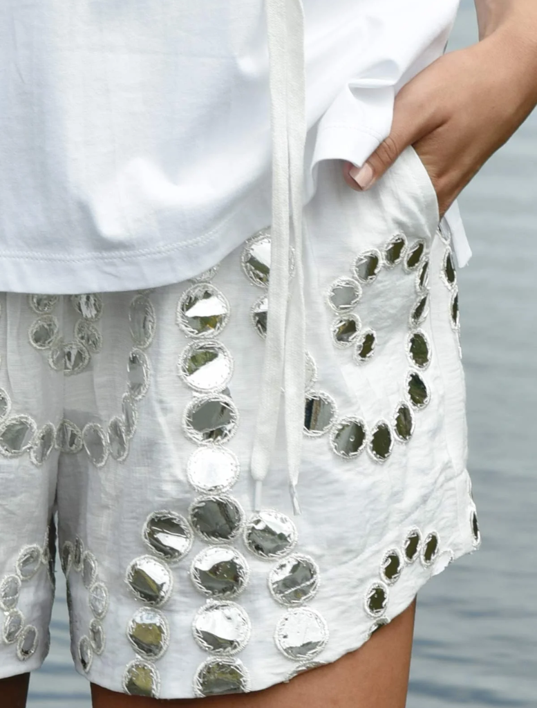 A woman in a white shirt and shorts stands by the water, reflecting on the serene environment around her.