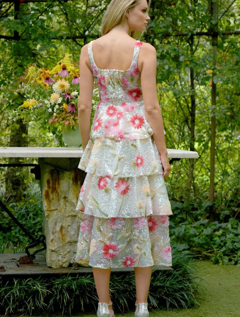 A woman in a floral dress stands before a garden, featuring an array of vibrant flowers and lush greenery.